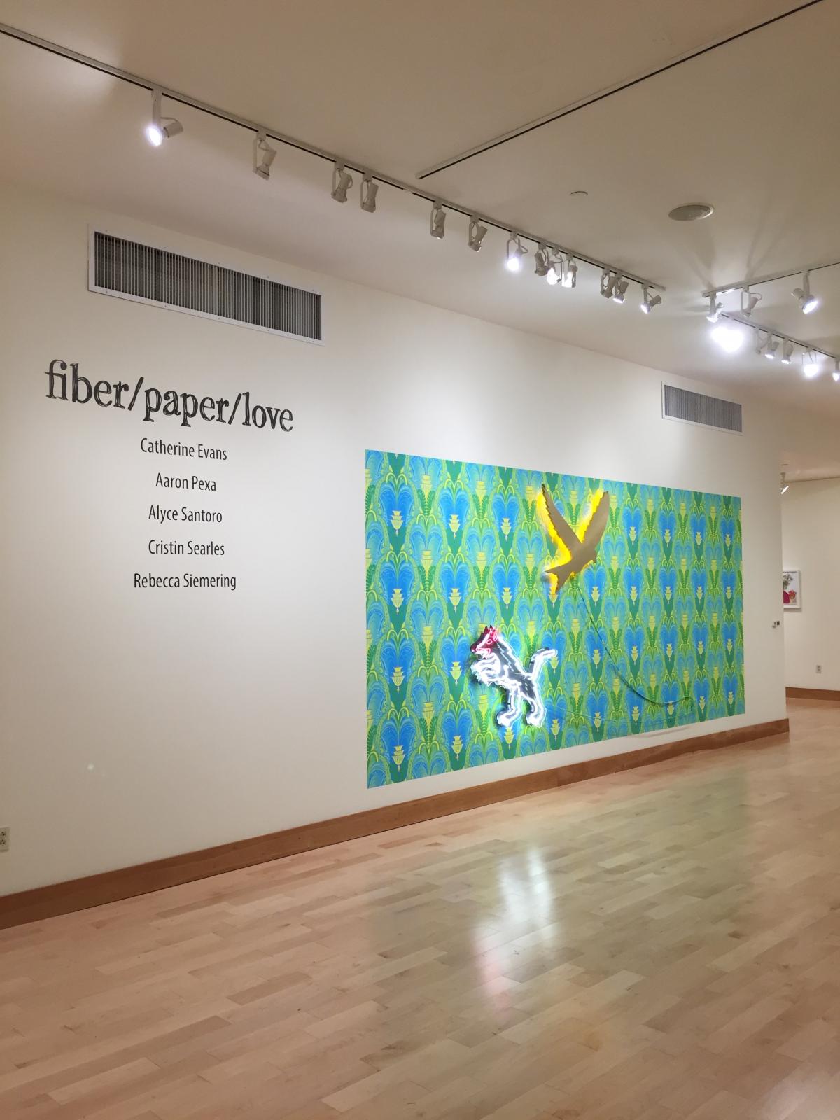 Gallery room view introductory text on the wall and Aaron Pexa’s “Garden Party…” piece
